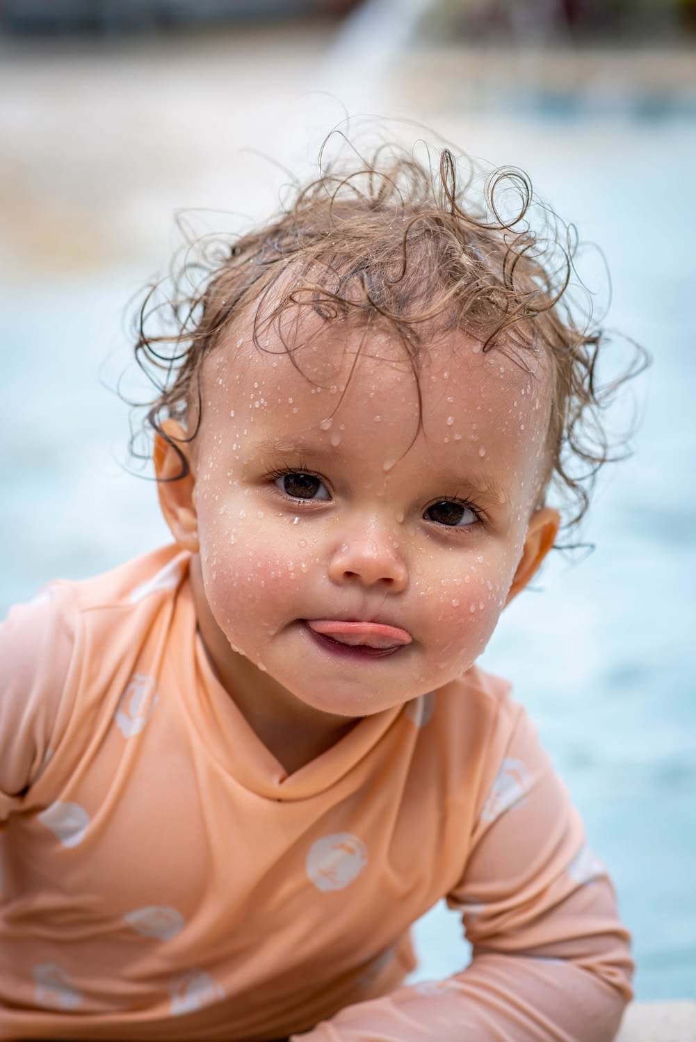 A baby girl with her tongue out and her wet from the pool.
