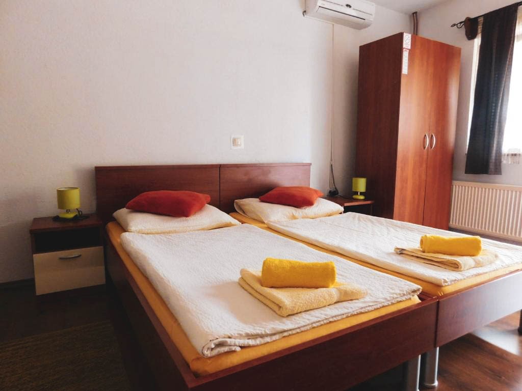 A king size bed with yellow covers and towels folded on them. It also has red pillows and a small brown table next to the bed, as well as a big closet on the other side.