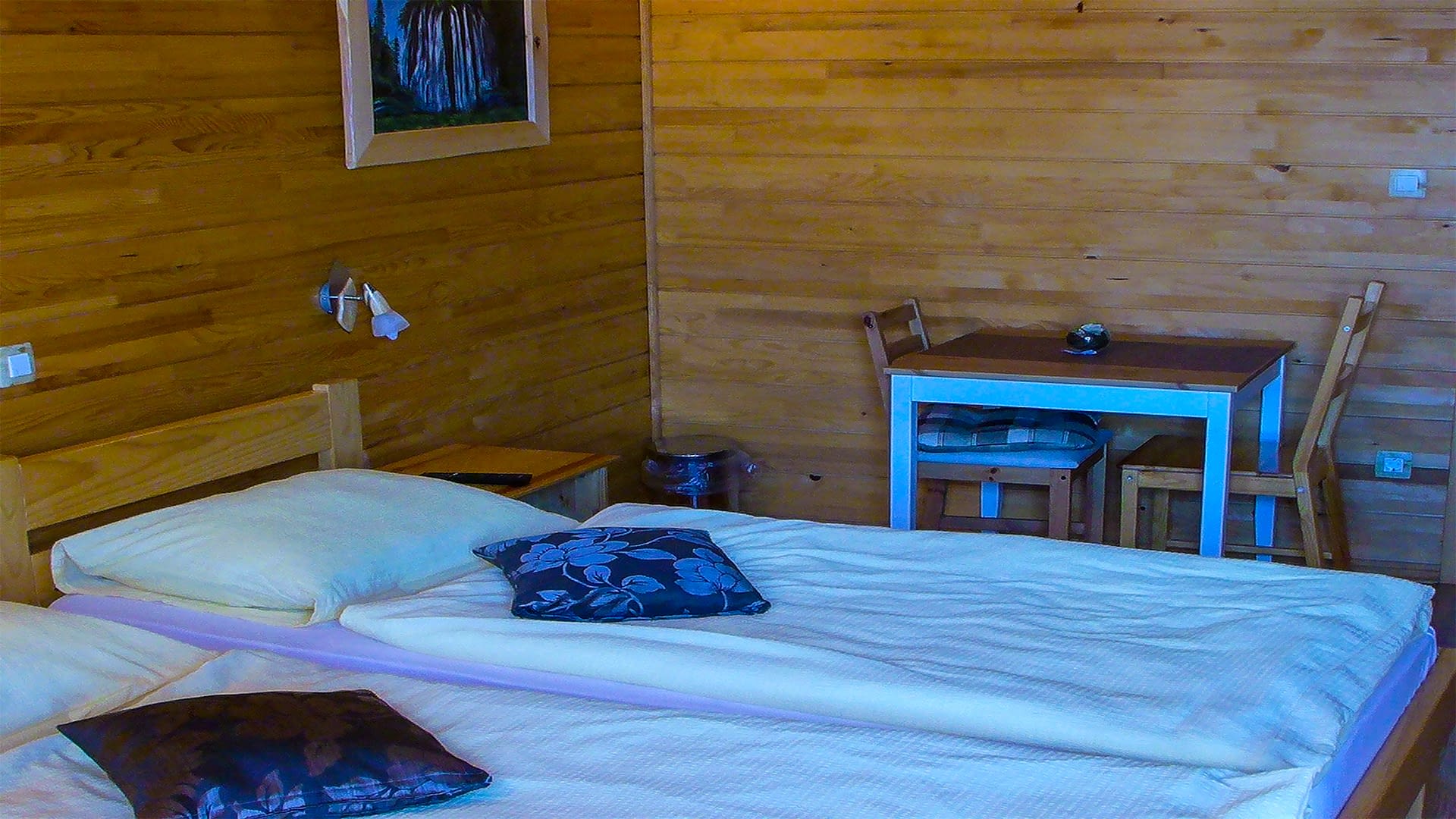 Two bed side by side with blue pillows on top of them. The walls in this room are made out of wood with a blue table sat beside them.