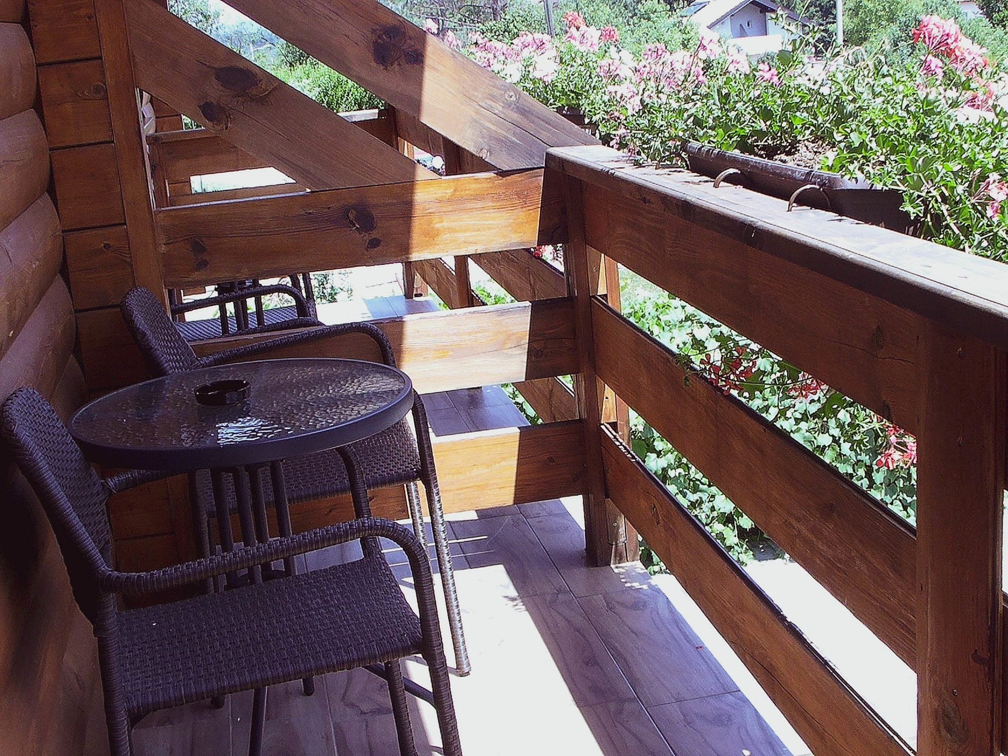 A wooden balcony from the deluxe room with flowers and two wooden chairs put next to a little wooden table.