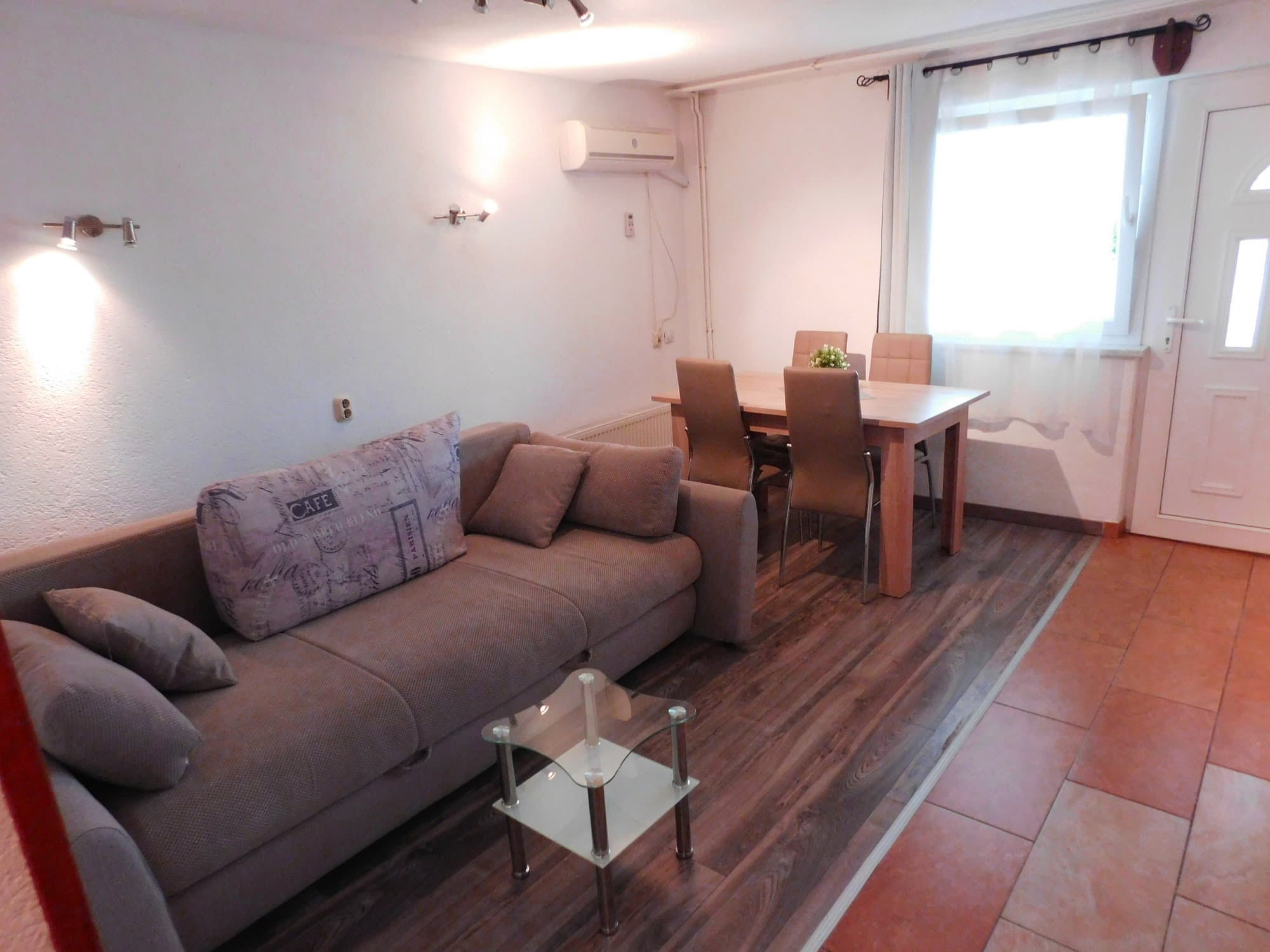 A wooden floor with a brown sofa on with a glass table in front of it. There is also a gray table in the background with 4 gray chairs that go with it.
