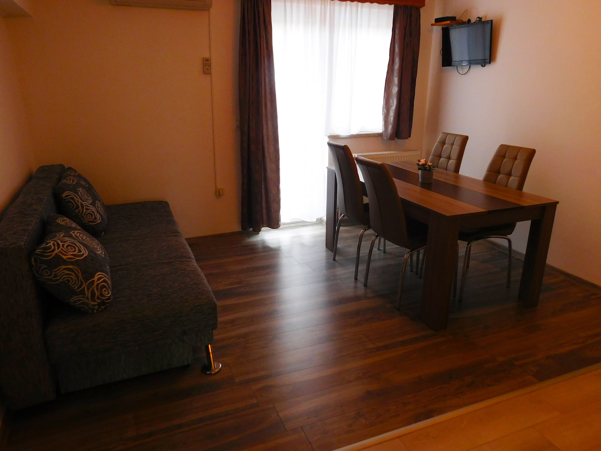 A wooden floor with a sofa bed on it. There is a brown table with 4 chairs and a TV set on the wool behind it.