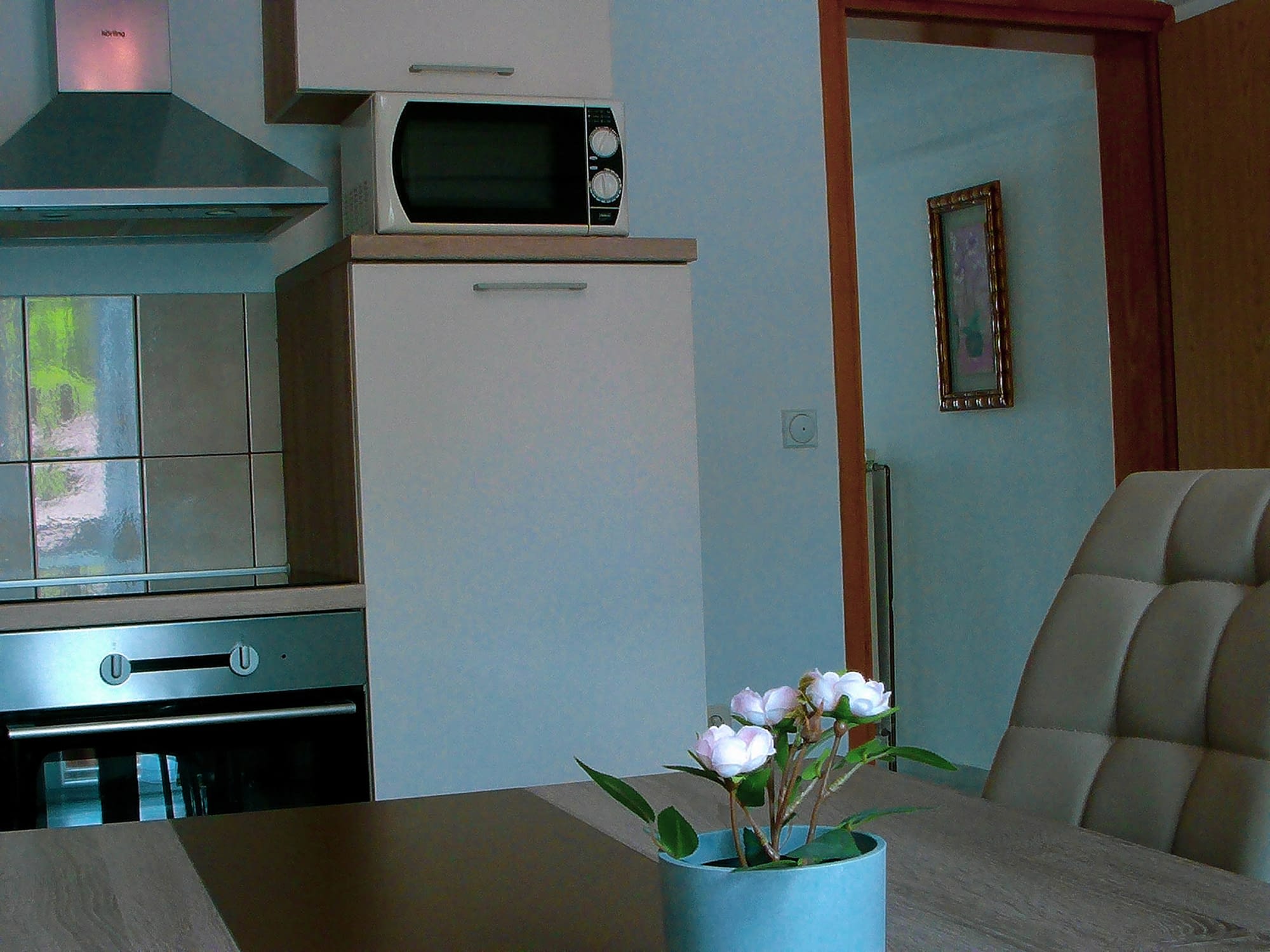 A white kitchen with a microwave, fridge and oven in it. There is also a big gray table in the middle with 4 gray chairs around it.