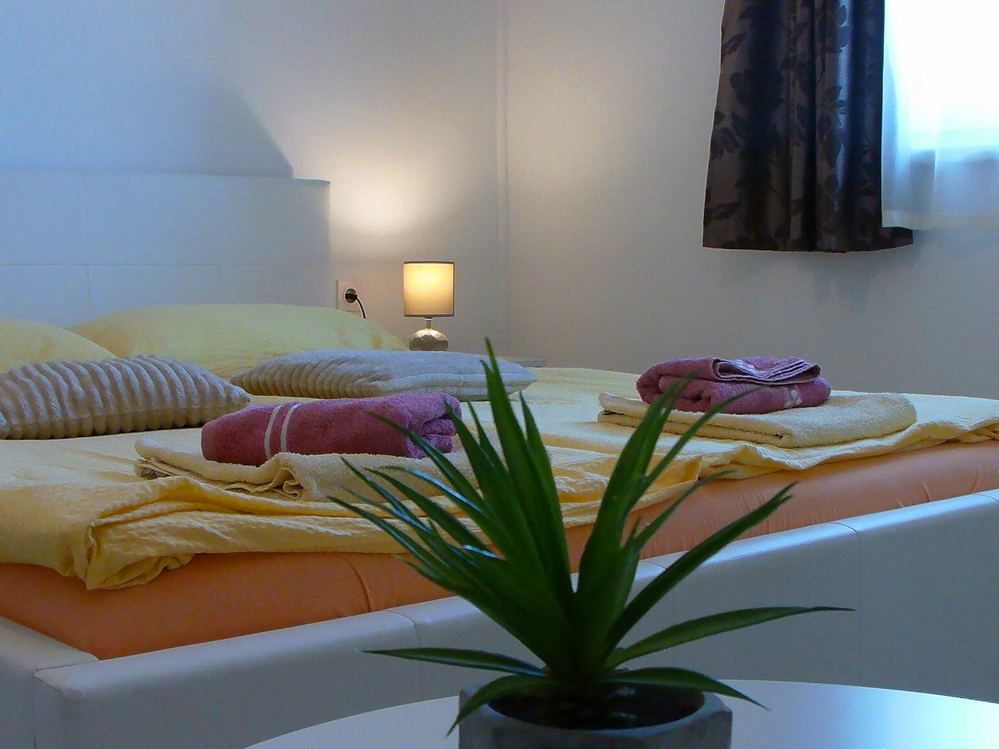 King size bed with orange covers and yellow pillows. There is also a green plant in the front shot of the picture.